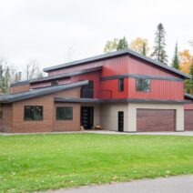 Klauer Steel Siding in brown and red on a home’s exterior. There are orange and green trees surrounding the home and a lush green lawn in the front area.